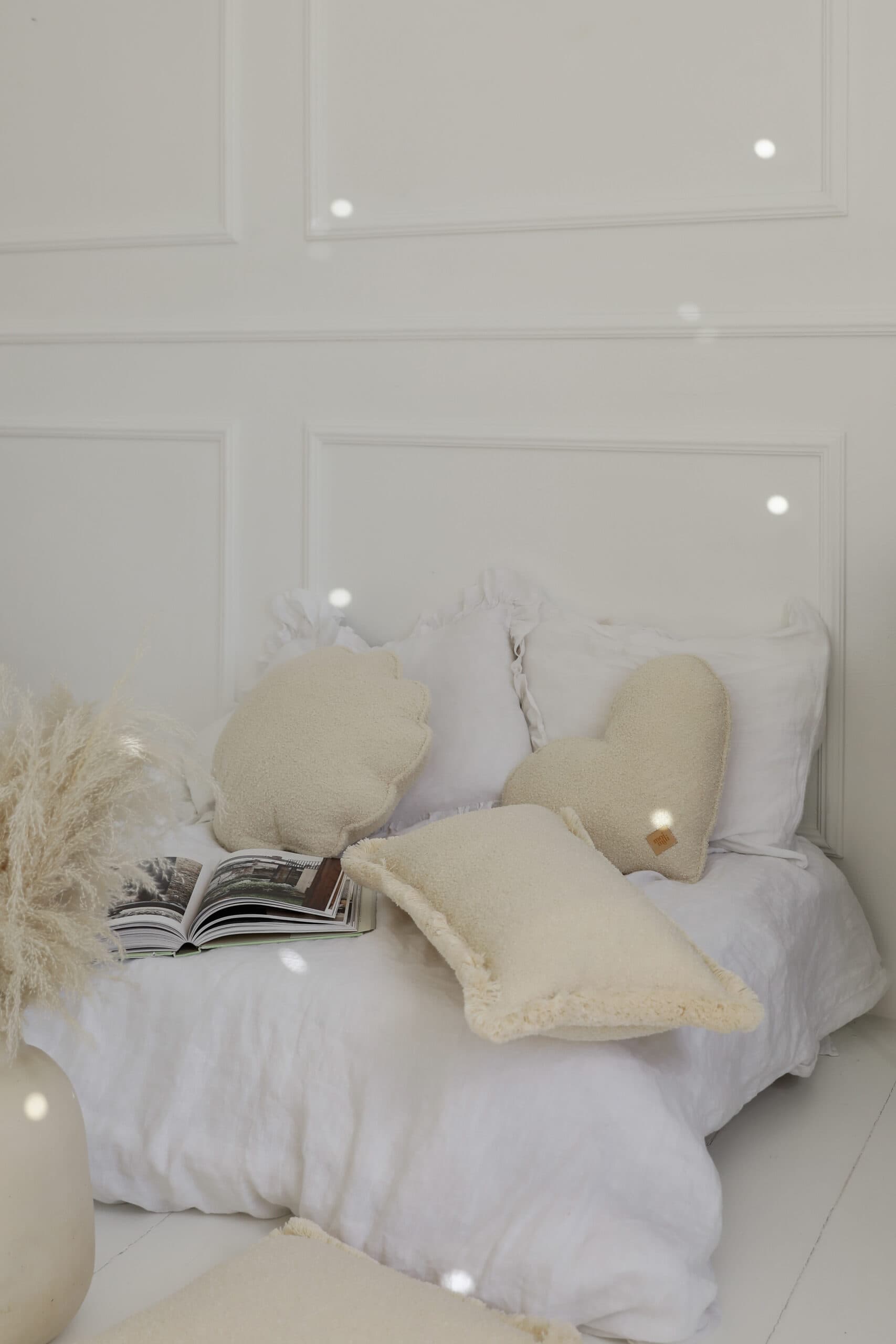 Przytulne, białe łóżko z miękkimi poduszkami w kształcie serca i muszli oraz otwartą książką na tle jasnej ściany.