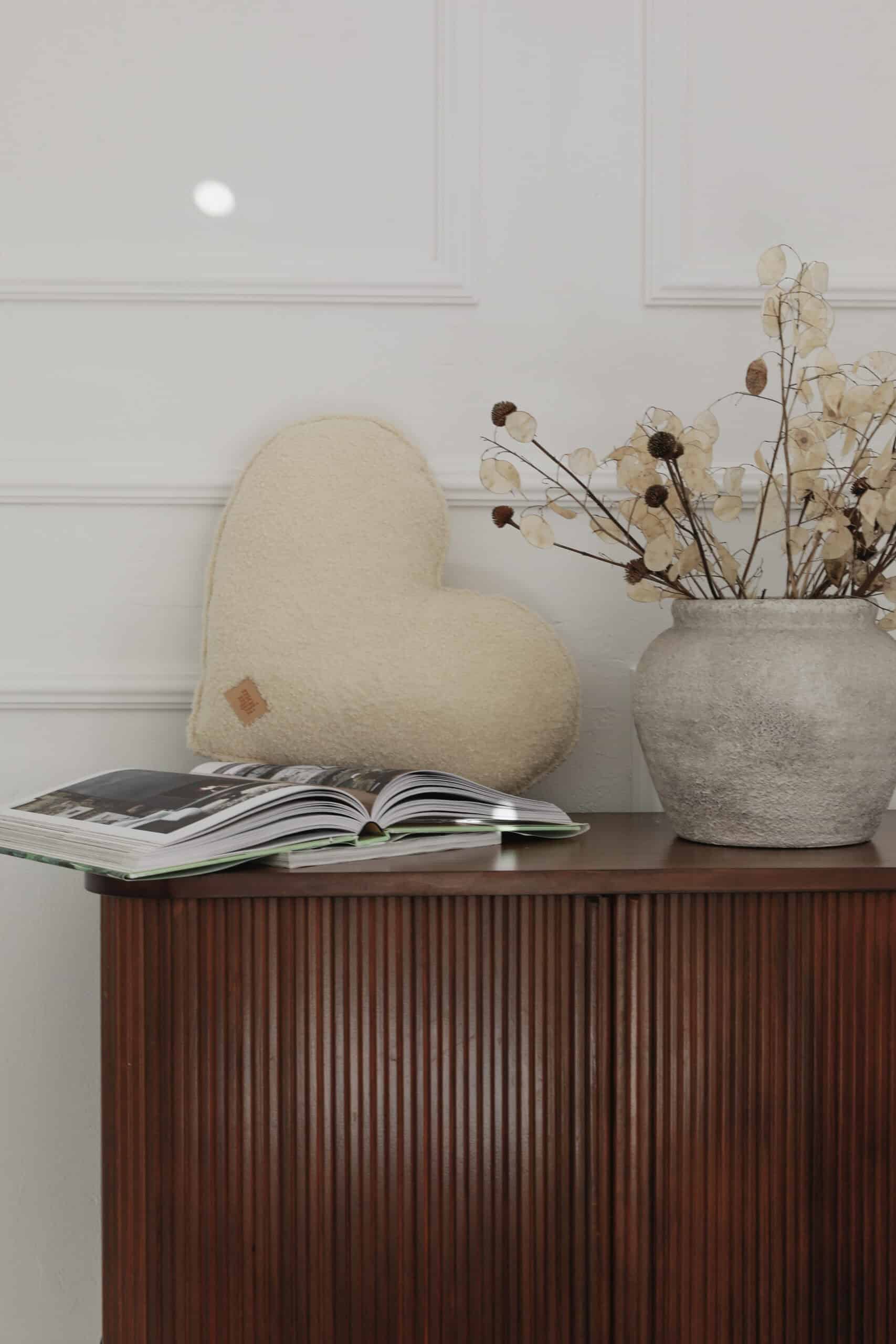 Drewniana komoda z otwartą książką, jasną poduszką w kształcie serca i donicą z suszonymi kwiatami.