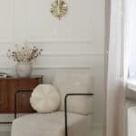 Nowoczesny fotel z jasnym obiciem i ozdobną poduszką obok drewnianej szafki i zasłony przy oknie