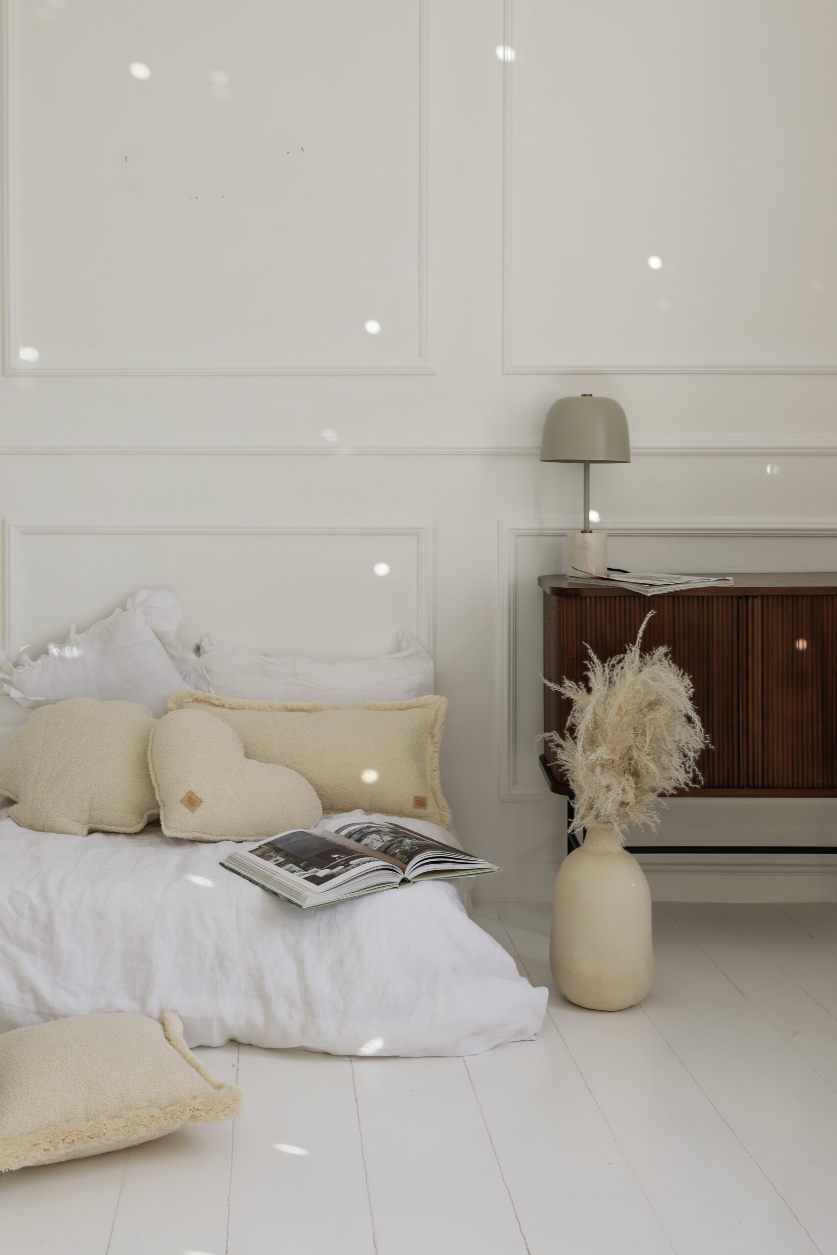 Przytulna sypialnia z białą pościelą, beżowymi poduszkami i wazonem z suszonymi roślinami obok drewnianej komody