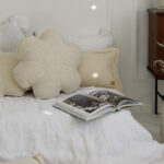 Przytulne, kremowe poduszki na białym łóżku z otwartą książką i drewnianą szafką obok