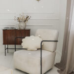 Beżowy fotel z czarnymi podłokietnikami i poduszką w kształcie muszli w jasnym wnętrzu z drewnianą szafką