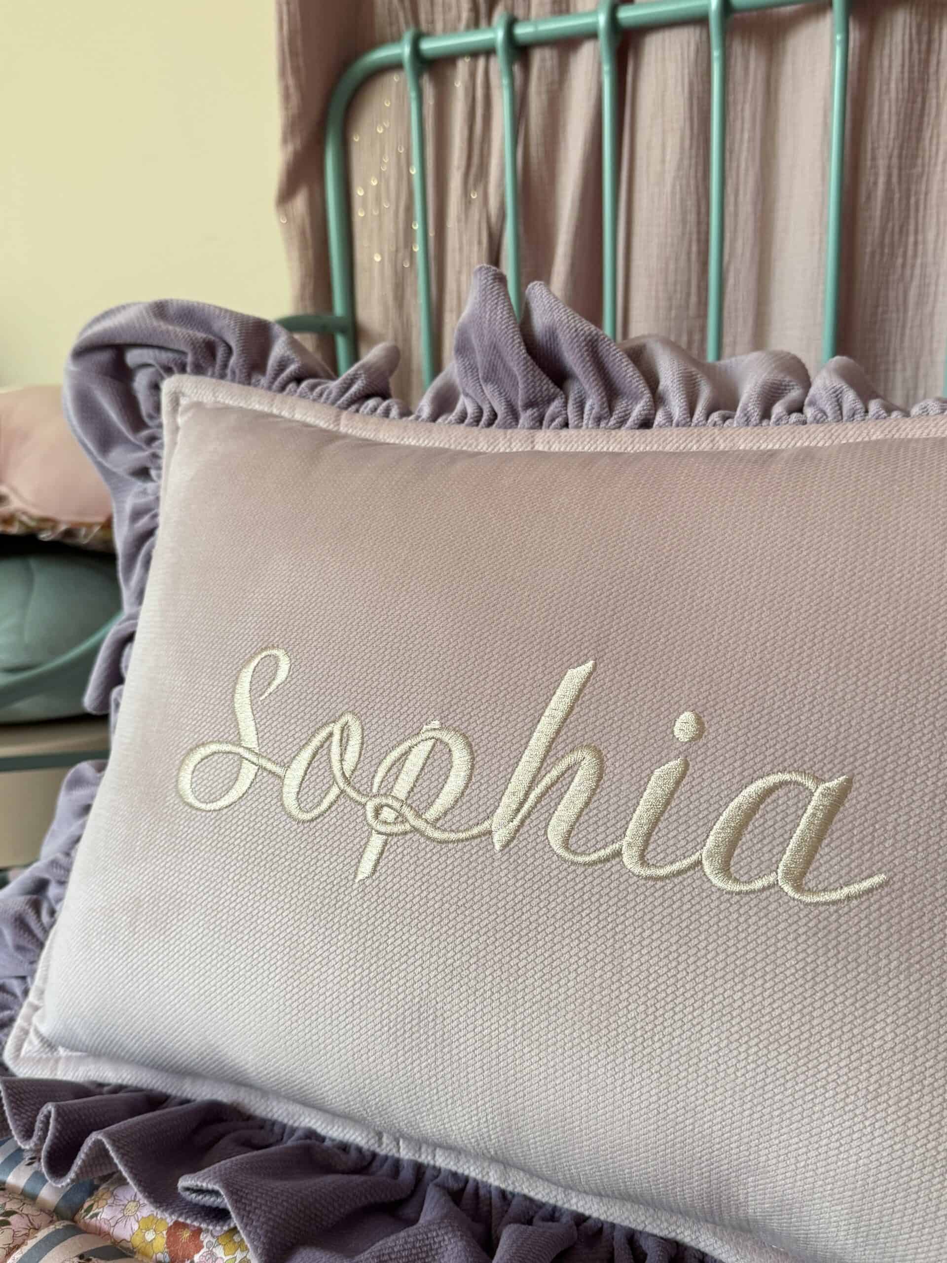 Różowa prostokątna poduszka z haftowanym imieniem Sophia na tle turkusowego stalowego zagłówka łóżka i jasnoróżowych zasłon.