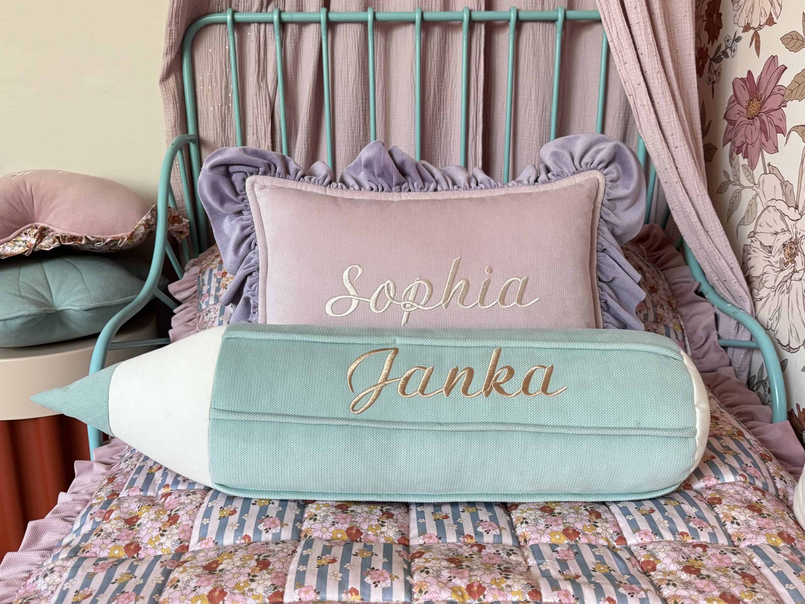 Łóżko dziecięce z turkusową metalową ramą, na którym znajdują się poduszki z imionami Sophia (różowa) i Janka (w kształcie ołówka, turkusowo-biała), na kolorowej narzucie w kwiaty i pasy.