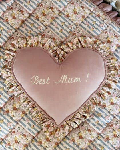 Różowa poduszka w kształcie serca z falbaną w kwiatowy wzór i napisem Best Mum! na pikowanym, patchworkowym tle w niebieskie paski i kwiaty.