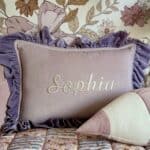 Różowa poduszka z falbankami w kolorze fioletowym, na której wyhaftowane jest imię Sophia, ustawiona na łóżku z kolorową narzutą oraz zukształtną poduszką w pasy, w tle tapeta z motywem kwiatowym w odcieniach brązu i bieli
