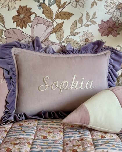 Różowa poduszka z falbankami w kolorze fioletowym, na której wyhaftowane jest imię Sophia, ustawiona na łóżku z kolorową narzutą oraz zukształtną poduszką w pasy, w tle tapeta z motywem kwiatowym w odcieniach brązu i bieli