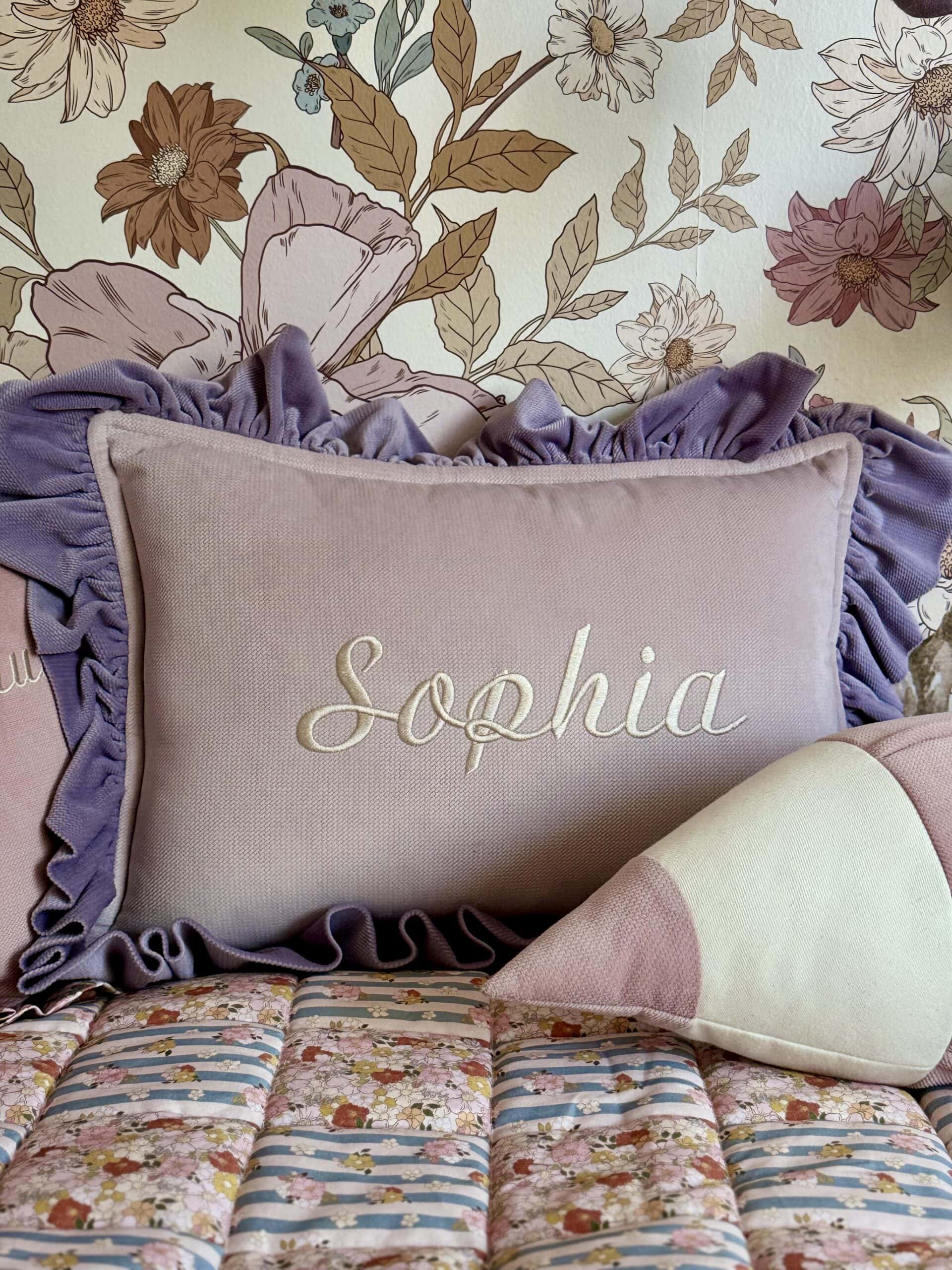 Różowa poduszka z falbankami w kolorze fioletowym, na której wyhaftowane jest imię Sophia, ustawiona na łóżku z kolorową narzutą oraz zukształtną poduszką w pasy, w tle tapeta z motywem kwiatowym w odcieniach brązu i bieli