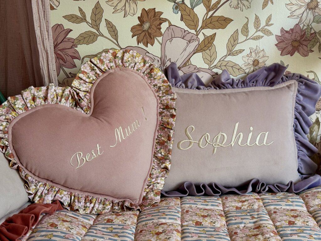 Dwa ozdobne poduszki na łóżku z kwiatowym wzorem, jedna w kształcie serca z napisem Best Mum!, druga prostokątna z napisem Sophia, obie otoczone falbankami, w tle tapeta z dużymi kwiatami.