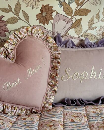 Dwa ozdobne poduszki na łóżku z kwiatowym wzorem, jedna w kształcie serca z napisem Best Mum!, druga prostokątna z napisem Sophia, obie otoczone falbankami, w tle tapeta z dużymi kwiatami.