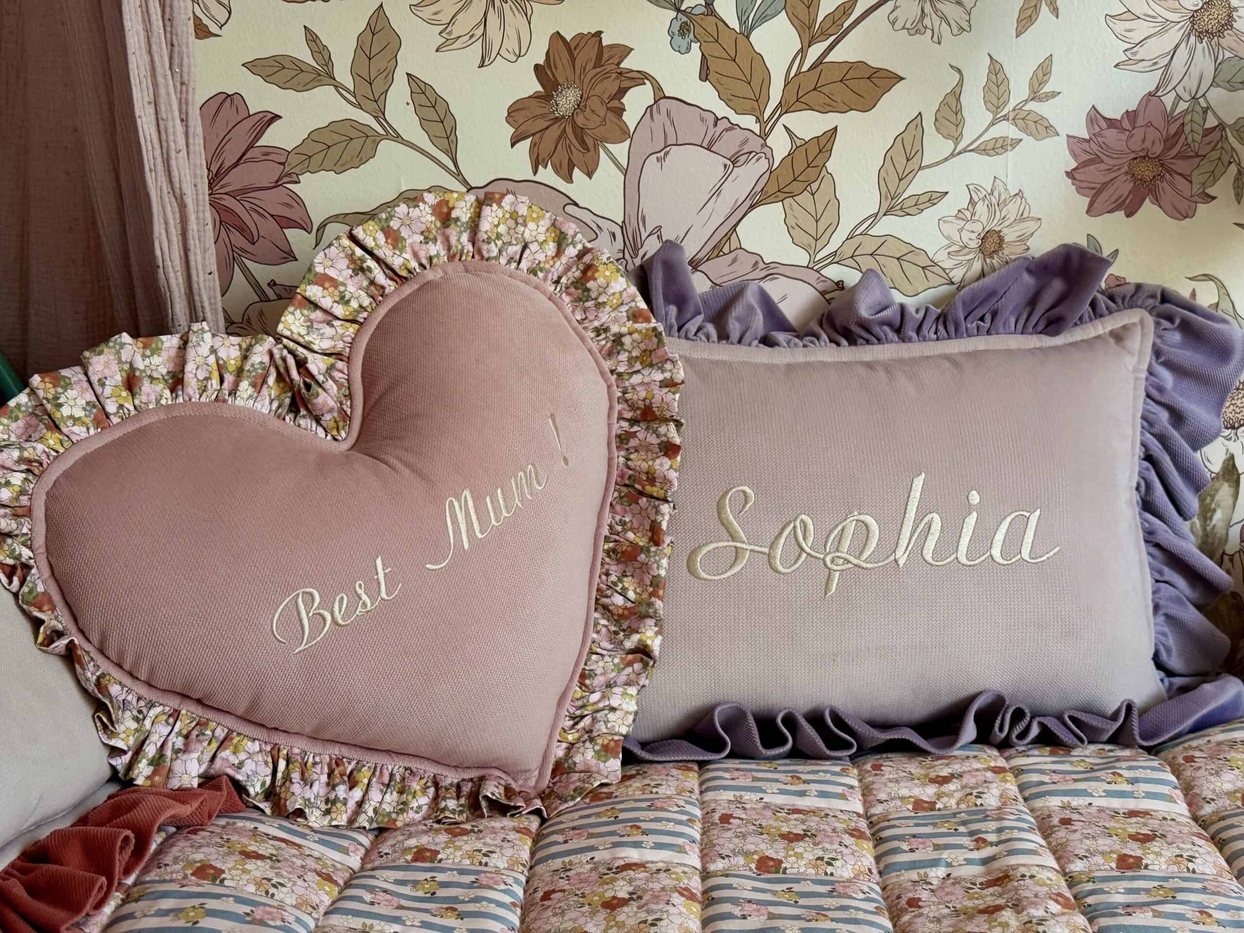 Dwa ozdobne poduszki na łóżku z kwiatowym wzorem, jedna w kształcie serca z napisem Best Mum!, druga prostokątna z napisem Sophia, obie otoczone falbankami, w tle tapeta z dużymi kwiatami.