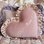 Różowa poduszka w kształcie serca z falbaną w kwiatowy wzór i napisem Best Mum! na tle innych poduszek z falbanami oraz kwiatowej tapety ściennej