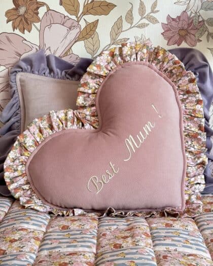 Różowa poduszka w kształcie serca z falbaną w kwiatowy wzór i napisem Best Mum! na tle innych poduszek z falbanami oraz kwiatowej tapety ściennej