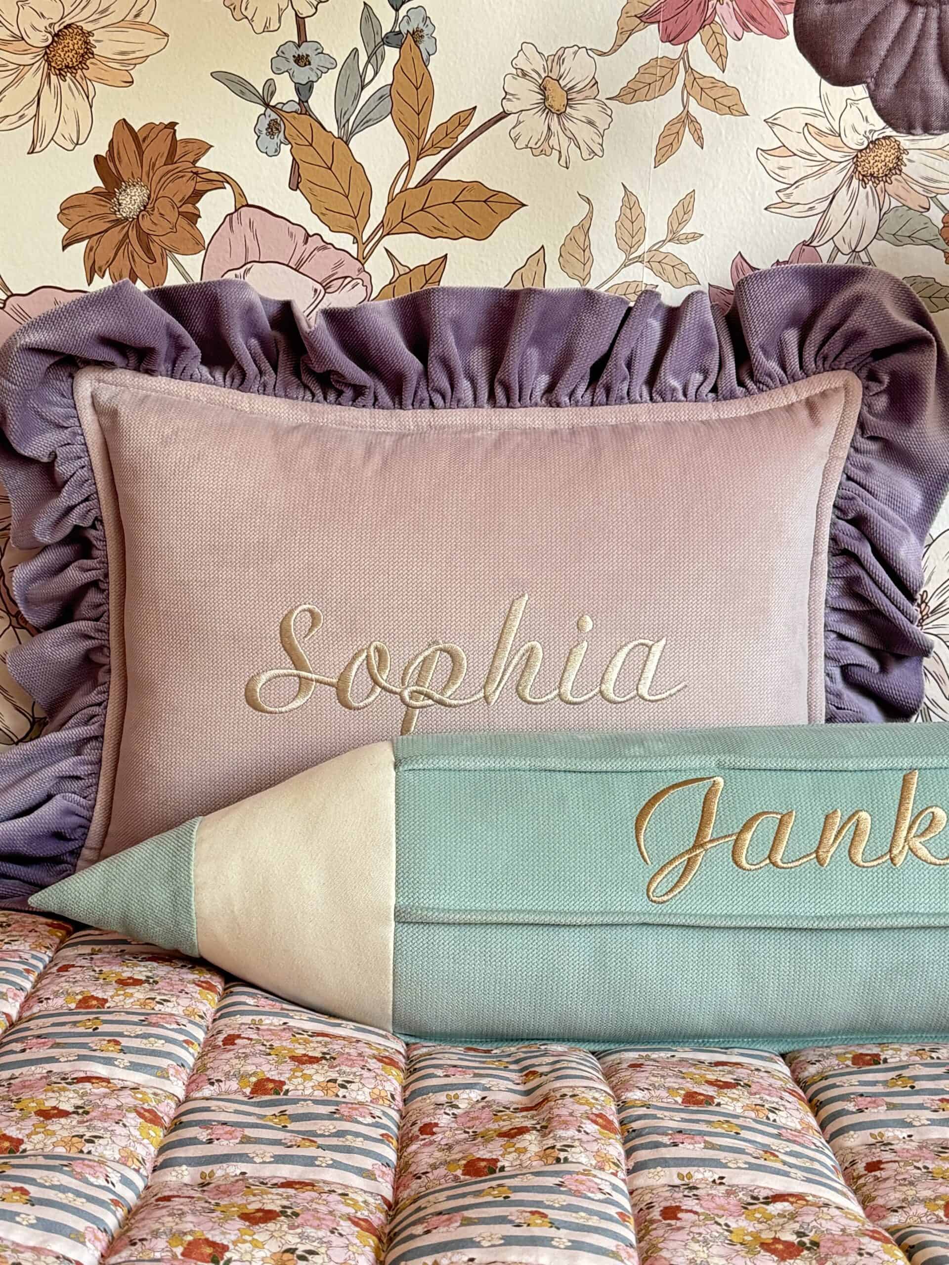 Różowa poduszka z falbanką i haftem imienia Sophia, zielono-biała poduszka w kształcie ołówka z haftem części imienia Janka na kolorowej narzucie, w tle tapeta z duży wzorem kwiatów w odcieniach beżu, brązu i różu