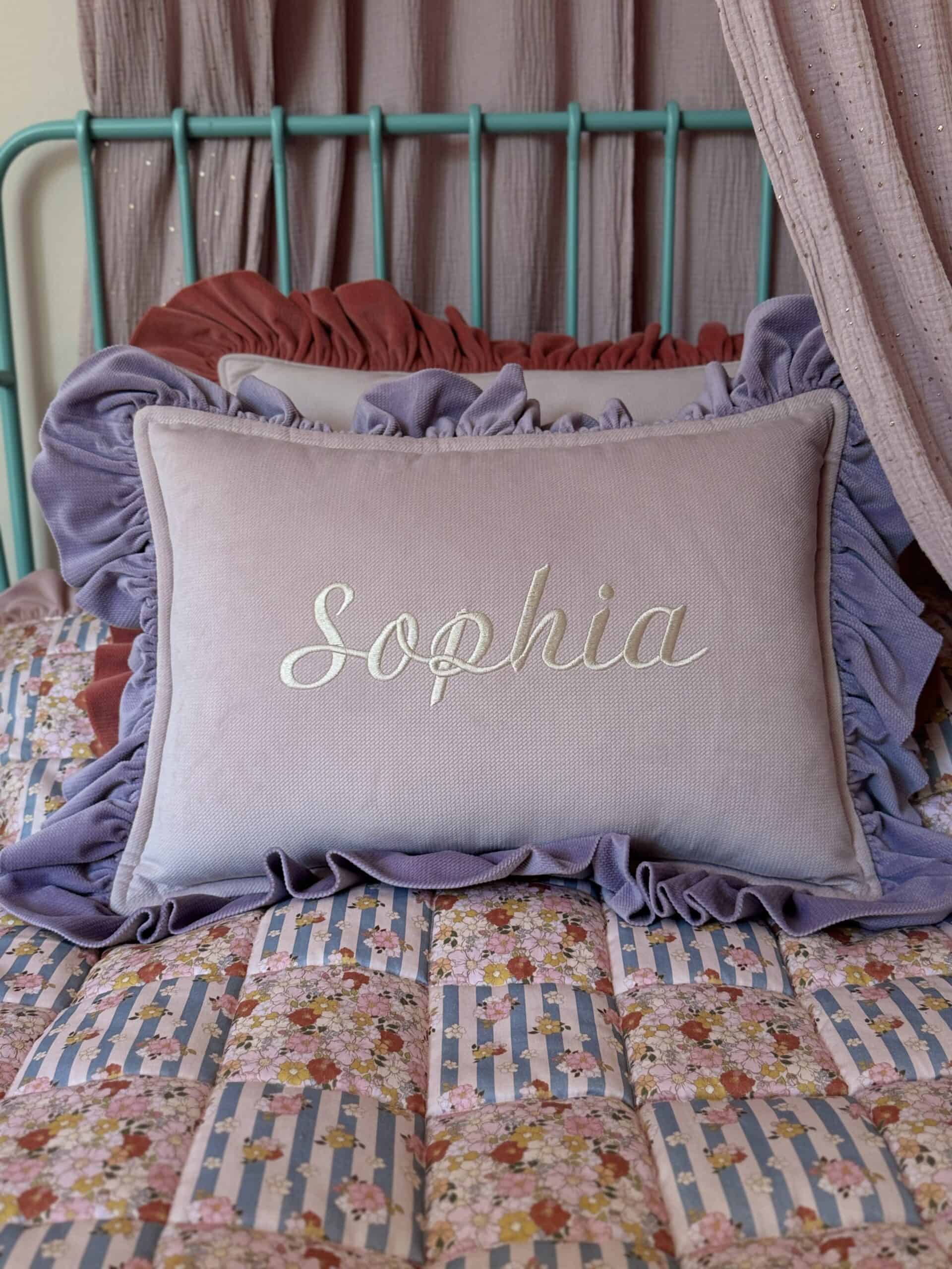Poduszka z falbankami w odcieniu lila z napisem Sophia na łóżku z patchworkową narzutą w niebieskie i różowe kwiaty, za nią druga poduszka w kolorze rdzy i metalowe niebieskie wezgłowie łóżka, w tle różowe zasłony.