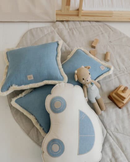 Trzy błękitne poduszki z beżową frędzlami i poduszka w kształcie samochodu na jasno szarym okrągłym dywanie z pluszową zabawką zwierzęcia i drewnianymi klockami, przy jasnym drewnianym łóżku na białej podłodze