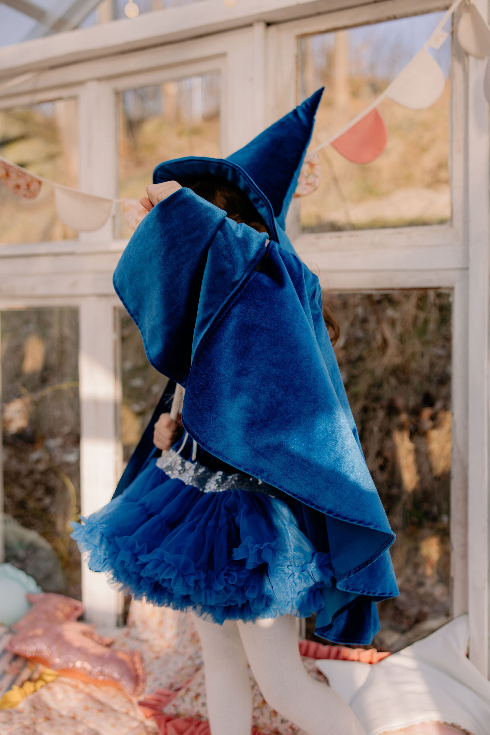 Dziecko w niebieskim kostiumie czarodzieja z peleryną i spódniczką tiulową, stojące bokiem w jasnym pomieszczeniu z dużymi oknami i girlandą kolorowych flag.