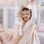 Mała dziewczynka w białej koszulce, beżowej pelerynie, cekinowej masce w kształcie kota i kapeluszu bawiąca się w jasnym, przytulnym pokoju udekorowanym girlandami i poduszkami.