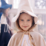 Uśmiechnięta dziewczynka w kremowym kapeluszu czarownicy i płaszczu, trzymająca błyszczącą złotą gwiazdę, z lekkim makijażem brokatu na twarzy, stoi w jasnym, rozmytym tle z inną osobą w niebieskim kostiumie czarownicy w tle.