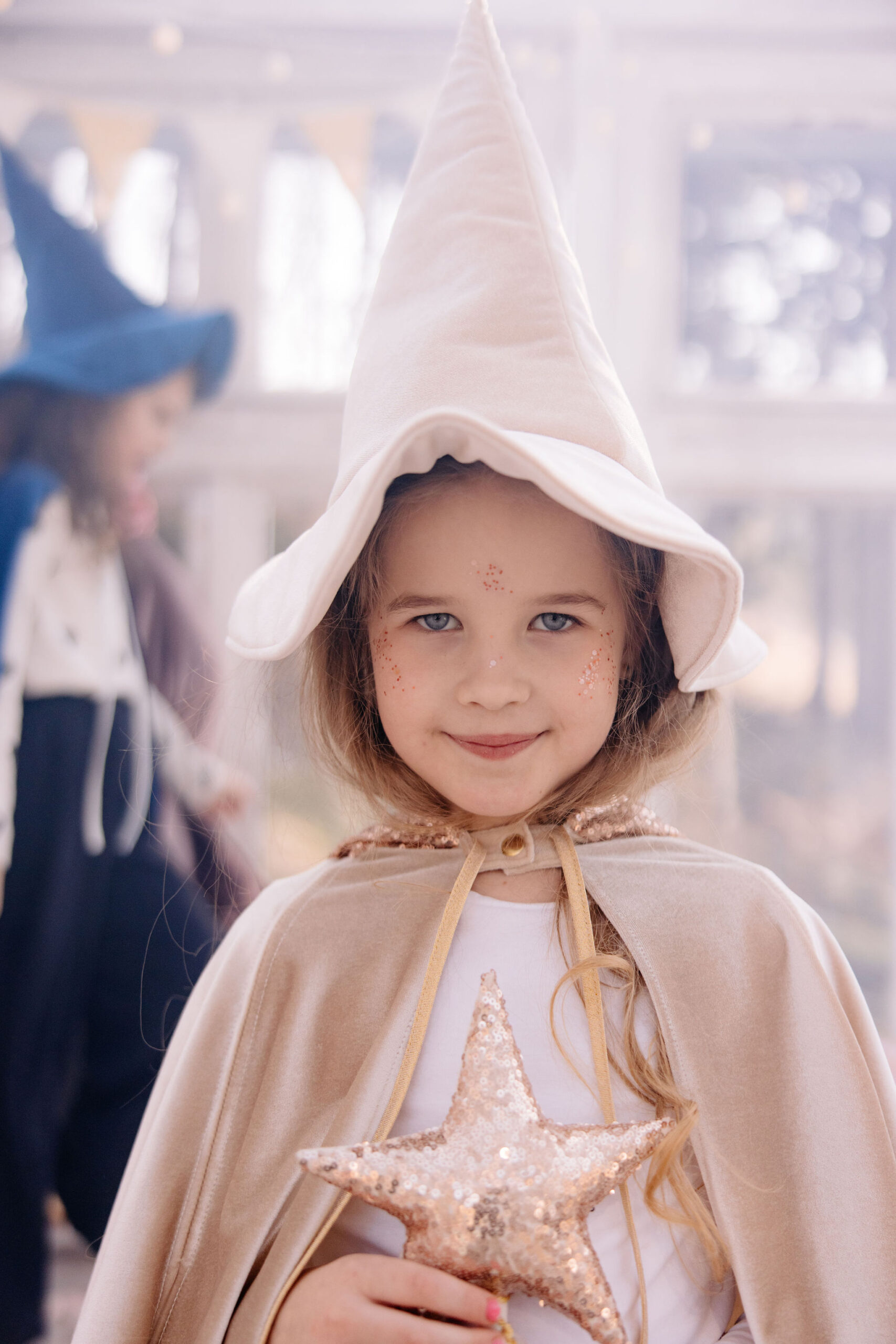 Uśmiechnięta dziewczynka w kremowym kapeluszu czarownicy i płaszczu, trzymająca błyszczącą złotą gwiazdę, z lekkim makijażem brokatu na twarzy, stoi w jasnym, rozmytym tle z inną osobą w niebieskim kostiumie czarownicy w tle.