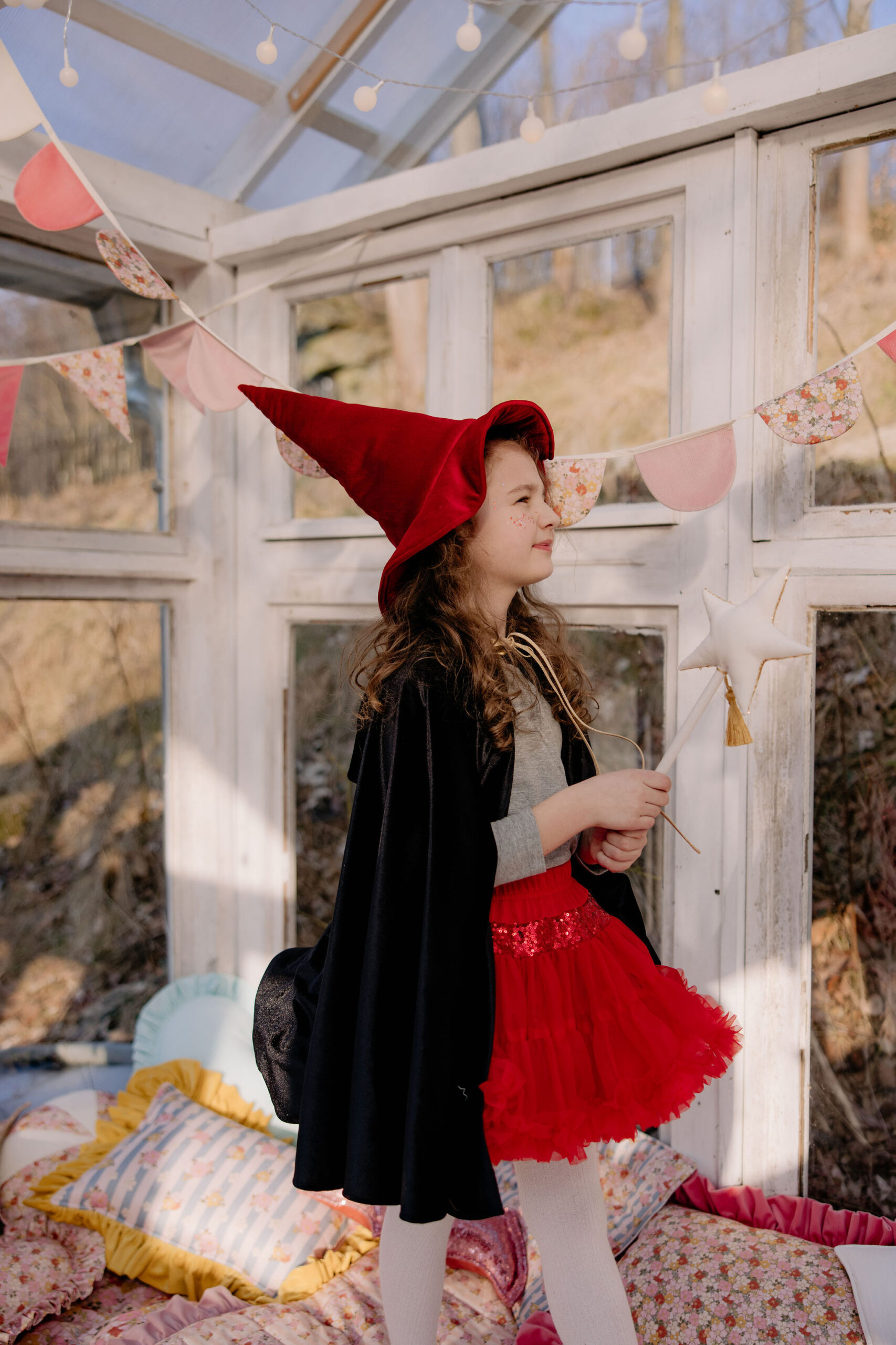 Dziewczynka w czerwonym kapeluszu czarownicy, czarnej pelerynie i czerwonej spódniczce, trzymająca różdżkę z gwiazdą, stojąca w jasnym, ozdobionym girlandami pomieszczeniu z poduszkami i miękkimi matami na podłodze.