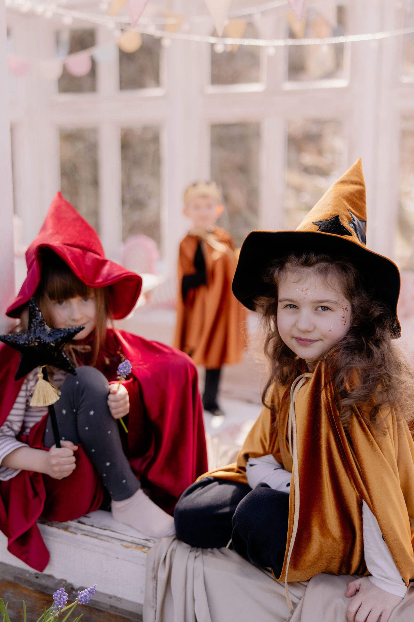 Troje dzieci ubranych w kostiumy czarownic i czarodziei w jasnym pokoju ozdobionym girlandami, dwie dziewczynki siedzą na podłodze w kolorowych pelerynach i kapeluszach, jedna trzyma czarną różdżkę i kwiatek, chłopiec stoi w tle w pomarańczowej pelerynie i złotej koronie.