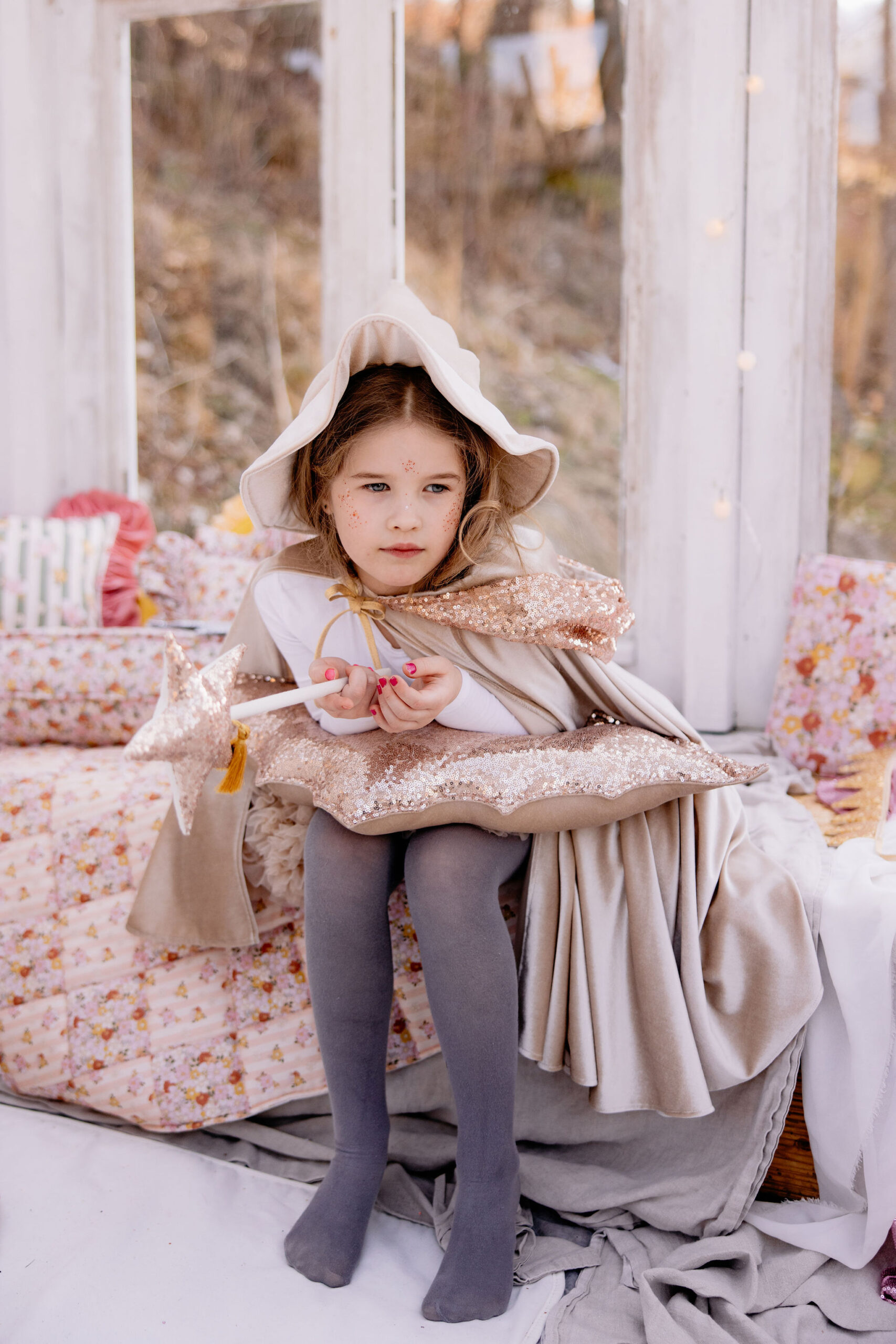 Dziewczynka w beżowym płaszczu z kapturem i szarych rajstopach siedzi na ławce, trzymając różdżkę w kształcie gwiazdy, z poduszką pokrytą cekinami na kolanach, w otoczeniu kwiecistych tkanin i białych ścian okna.