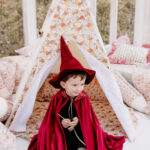 Mały chłopiec w czerwonym kapeluszu i pelerynie siedzi w namiocie w typie tipi w jasnym pomieszczeniu, otoczony poduszkami i dekoracjami w kwiatowy wzór.