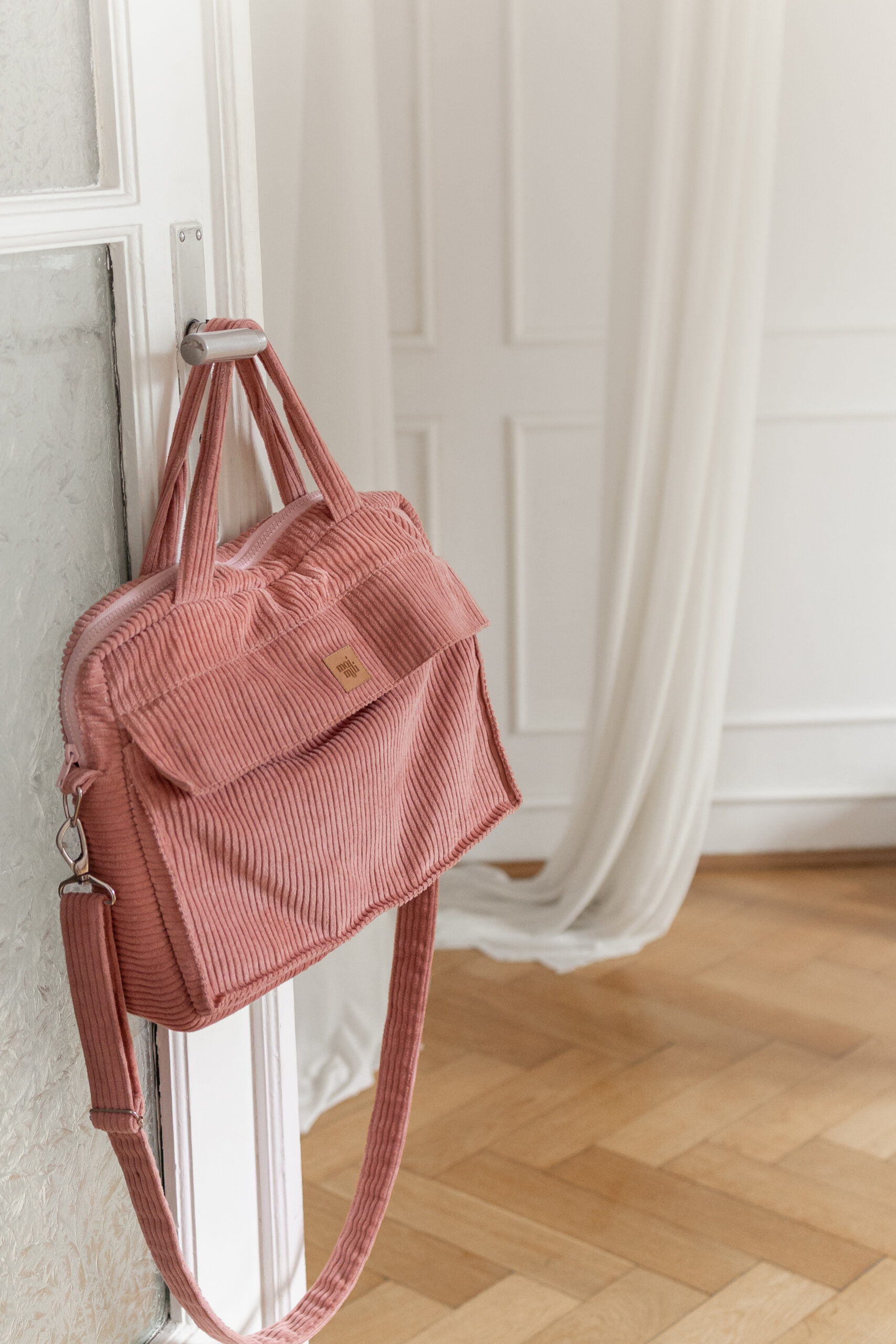 Różowa torba z prążkowanego materiału zawieszona na srebrnym uchwycie drzwi w jasnym wnętrzu z drewnianą podłogą i białą zasłoną w tle