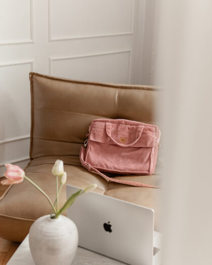 Różowa torba na beżowej skórzanej kanapie w pomieszczeniu z kremową ścianą, na pierwszym planie na marmurowym stoliku wazon z trzema tulipanami i laptop Apple obok.