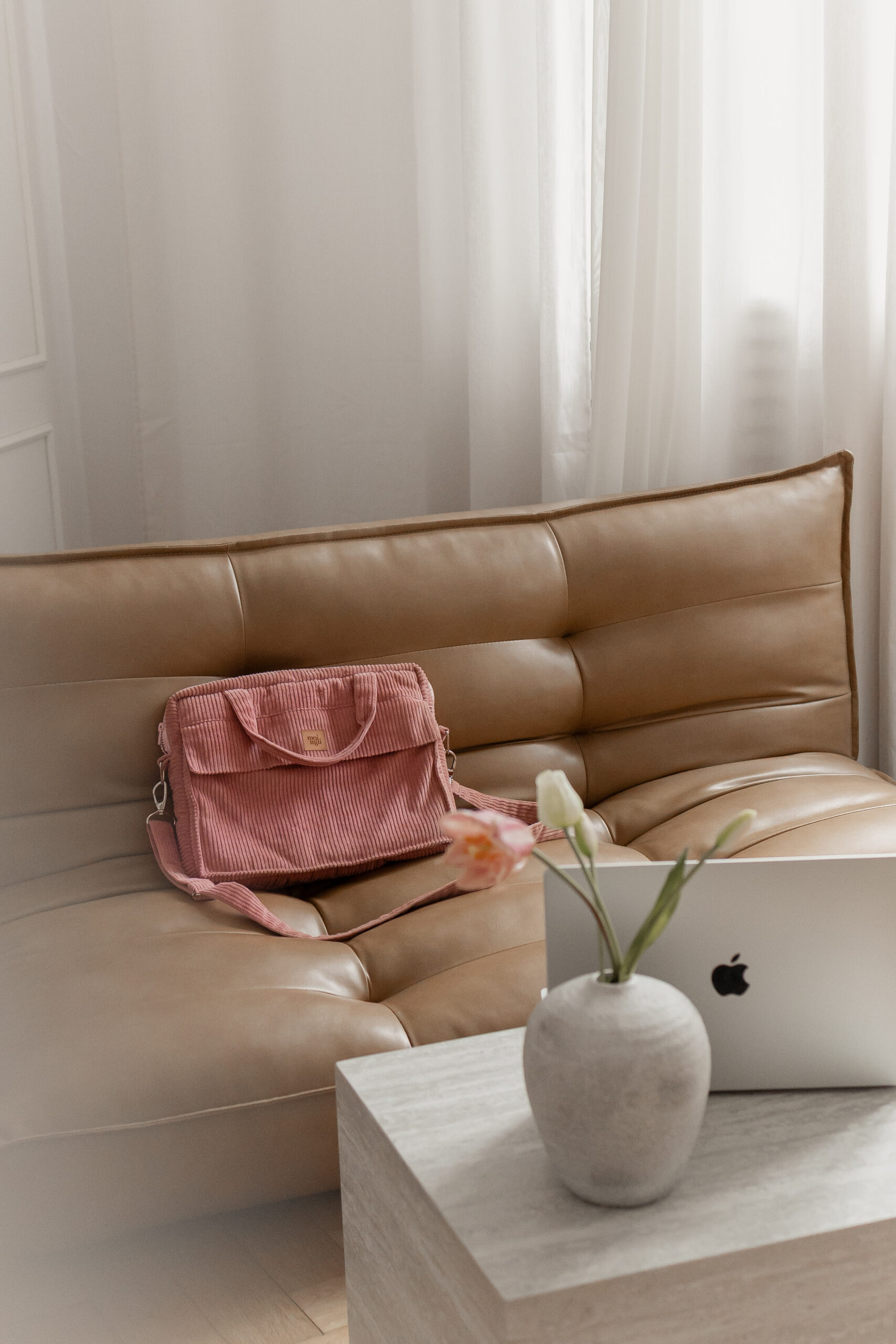 Brązowa skórzana sofa z różową torebką z teksturą na niej, obok stoi jasnoszary stolik z wazonem z różowymi i białymi kwiatami oraz srebrnym laptopem Apple, w tle białe zasłony.