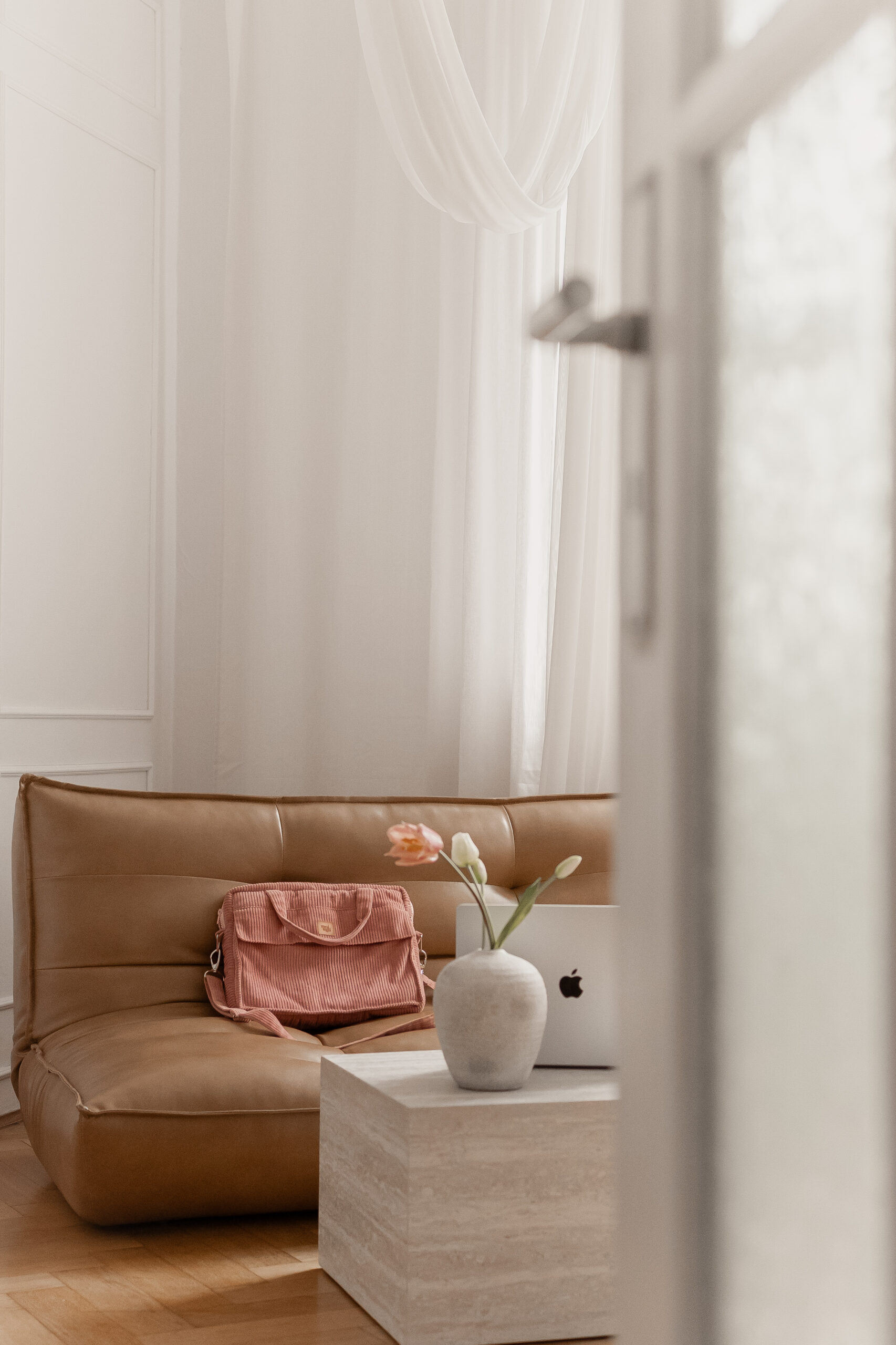 Brązowa skórzana sofa w jasnym wnętrzu, na której znajduje się różowa torba, obok stolik z jasnego kamienia z szarą wazonem i kwiatami oraz laptopem Apple.