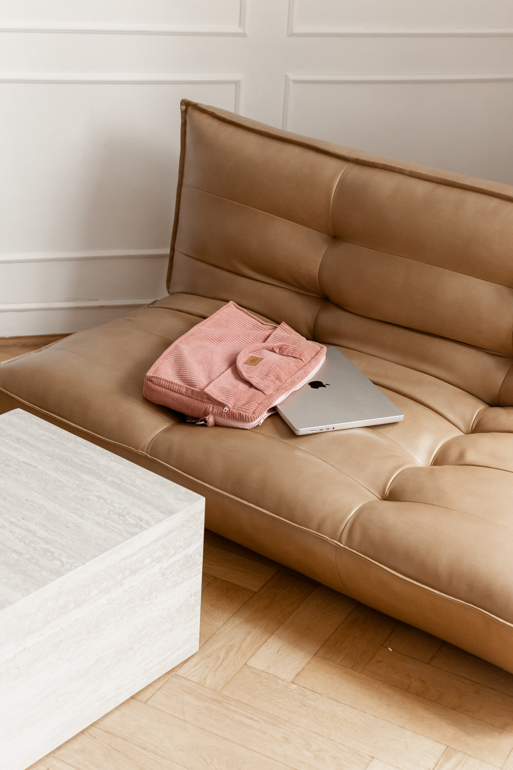 Beżowa skórzana sofa z zamkniętym srebrnym laptopem i różowym etui na laptop na siedzisku, obok biały marmurowy stolik na jasnej drewnianej podłodze w jasnym pokoju z białą boazerią na ścianie