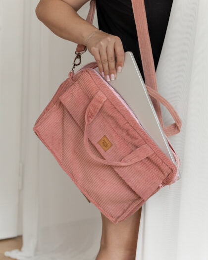 Osoba wkładająca srebrny laptop do różowej torby na ramię z materiału przypominającego sztruks, ubrana w czarną bluzkę i srebrną bransoletkę, stojąca przy białej ścianie i firance w jasnym pomieszczeniu.