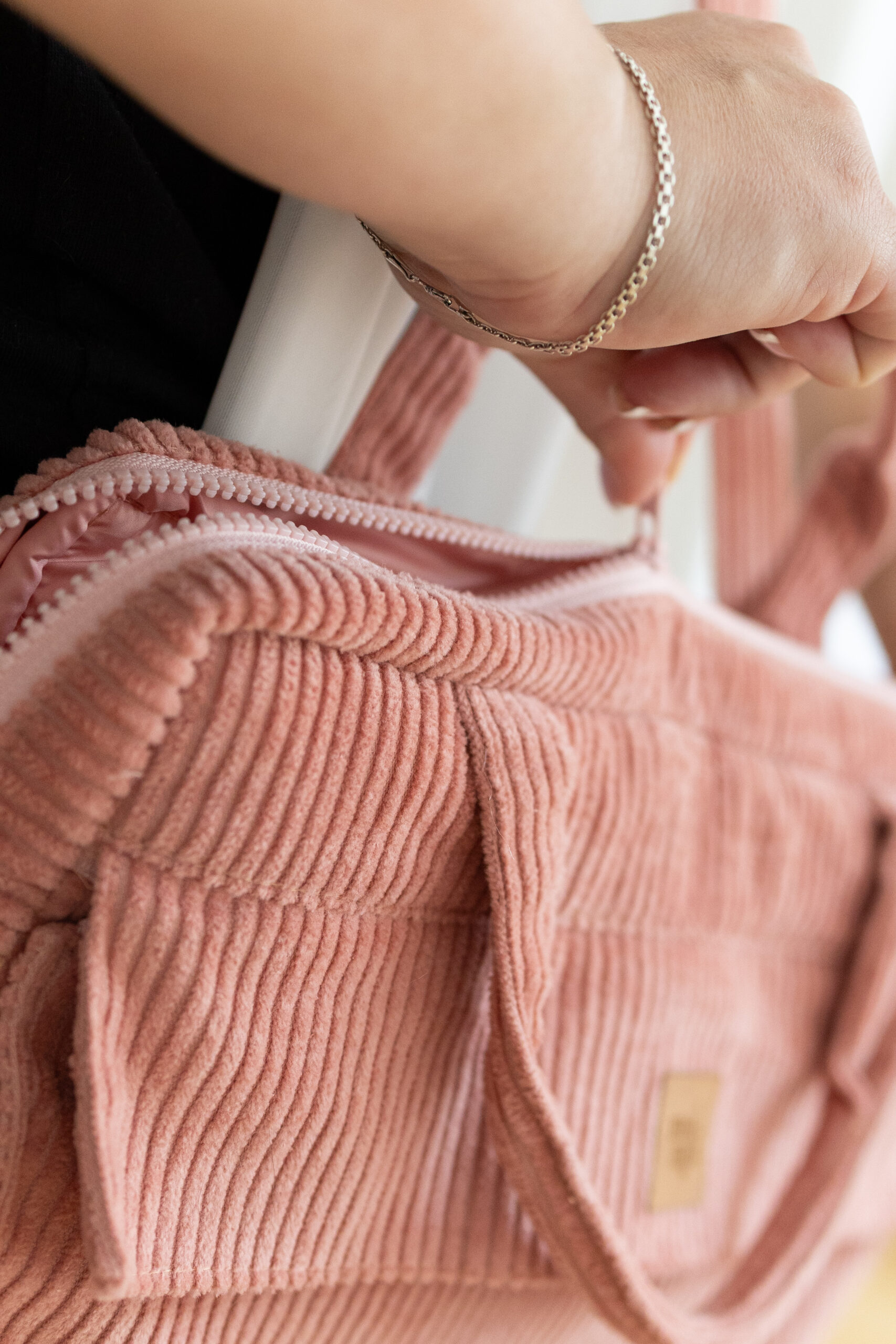 Ręka z srebrną bransoletką rozpinająca zamek błyskawiczny na różowej, prążkowanej torbie z tkaniny sztruksowej.