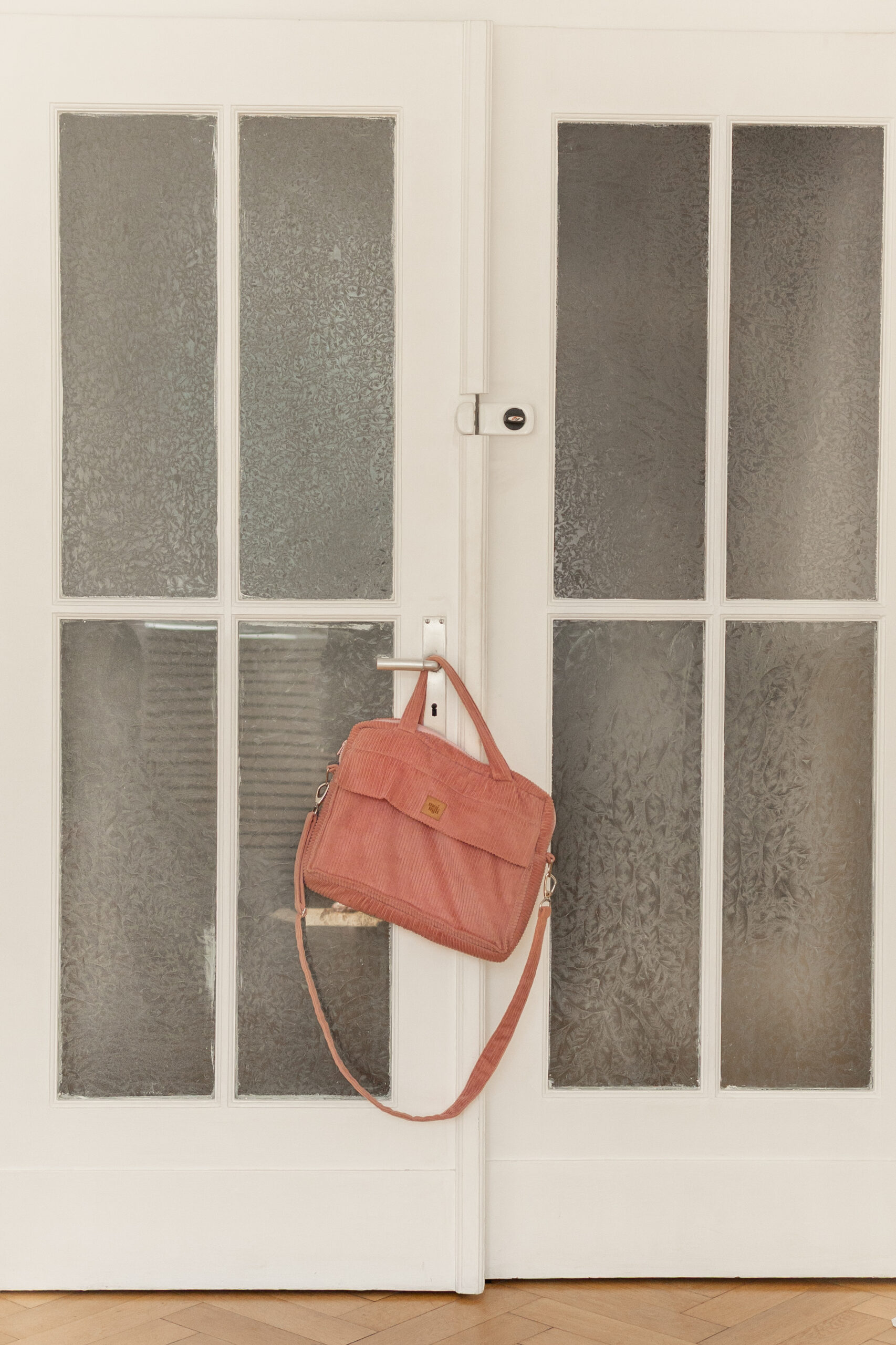 Różowa torba na ramię wisząca na białych drzwiach z matowymi szybami w stylu francuskim na tle drewnianej podłogi w jodełkę.