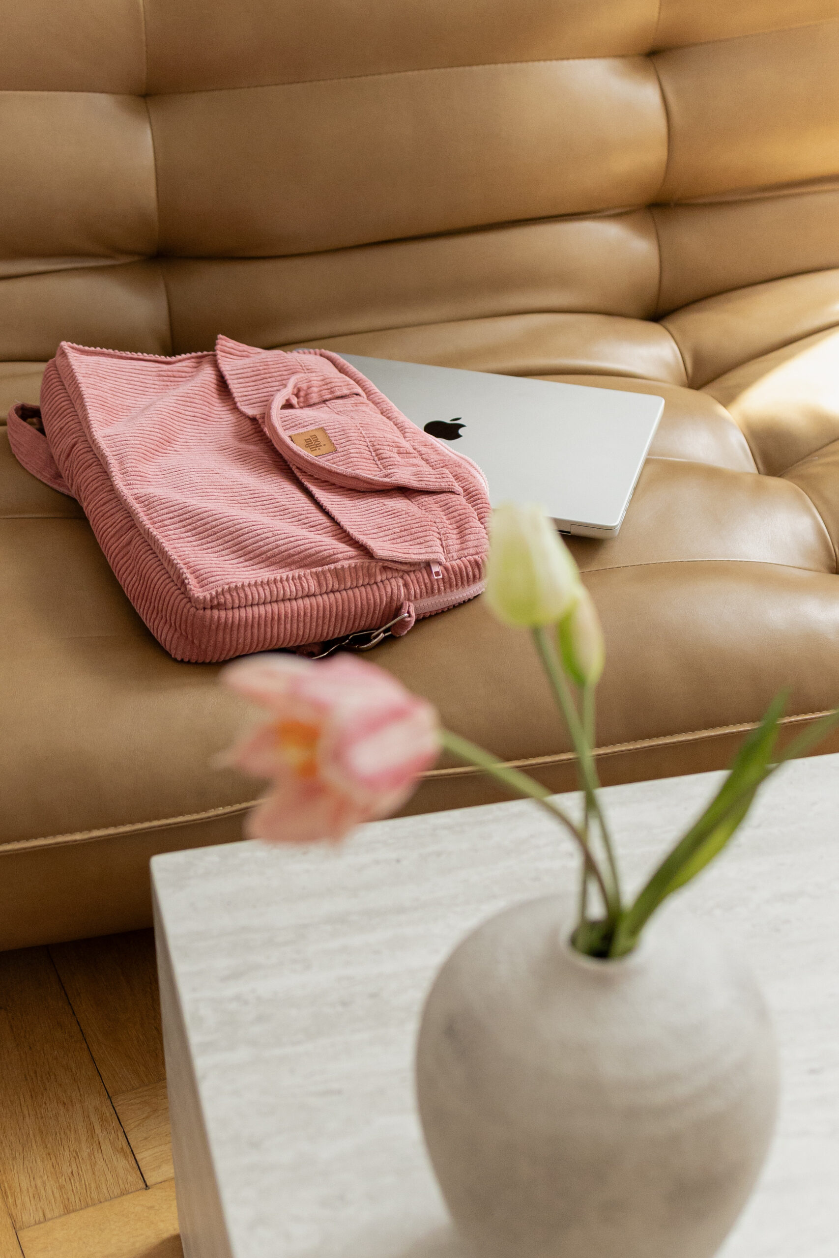 Torba na laptopa z różowego sztruksu i srebrny laptop na beżowej skórzanej kanapie z rozmytym wazonem z kwiatami na pierwszym planie na jasnym stole