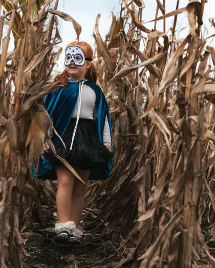 Dziewczynka w niebieskiej pelerynie i białej masce na twarzy stoi między suchymi kolbami kukurydzy na polu.
