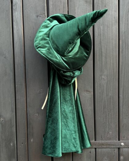 Zielony aksamitny kapelusz czarownicy z szerokim rondem i wysokim, zakrzywionym szpicem, wiszący na drewnianych deskach drzwi.