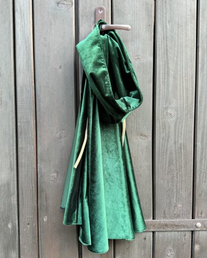 Zielony aksamitny fartuch z kapturem wiszący na brązowych drewnianych drzwiach na metalowym haczyku.