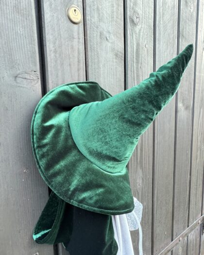 Zielony aksamitny kapelusz czarownicy zawieszony na drewnianych, szarych deskach z widocznym metalowym zamkiem.