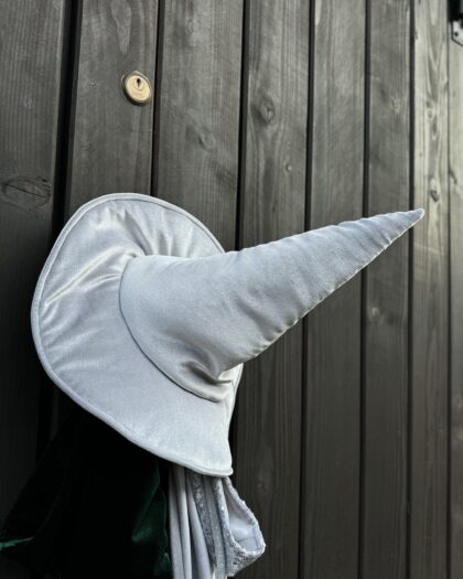 Biały kapelusz czarodzieja z długim, spiczastym zakończeniem zawieszony na ciemnych drewnianych drzwiach z widocznym złotym zamkiem.