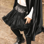 Fragment sylwetki dziecka w czarnej tiulowej spódnicy z cekinami, czarnych rajstopach, czarnych lakierowanych botkach i w czarnej pelerynie, stojącego na ziemi z lekko ugiętymi nogami.