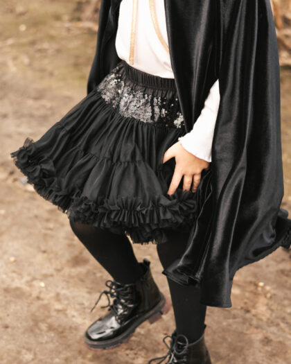 Fragment sylwetki dziecka w czarnej tiulowej spódnicy z cekinami, czarnych rajstopach, czarnych lakierowanych botkach i w czarnej pelerynie, stojącego na ziemi z lekko ugiętymi nogami.