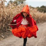 Dziewczynka w czerwonym pelerynie z kapturem i pomarańczowej spódnicy z falbanek stoi na polnej drodze obok suchego kukurydzianego pola, patrząc w dół i trzymając spódnicę.