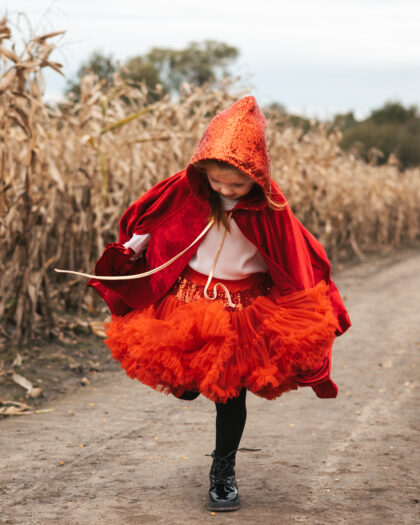 Dziewczynka w czerwonym pelerynie z kapturem i pomarańczowej spódnicy z falbanek stoi na polnej drodze obok suchego kukurydzianego pola, patrząc w dół i trzymając spódnicę.