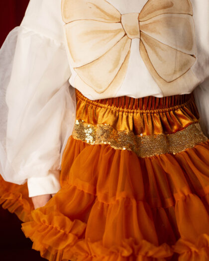 Fragment dziecięcego stroju z białą bluzką ozdobioną dużą beżową kokardą oraz pomarańczową spódnicą z falbanami i błyszczącym paskiem z cekinów.