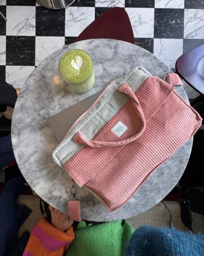 Okrągły marmurowy stół z różowo-szarym plecakiem, laptopem oraz zieloną herbatą matcha z rysunkiem serca na piance, obok ręka ubrana w kolorowy, żywy sweter siedzącej osoby, widok z góry.