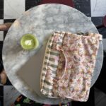 Okrągły marmurowy stolik na tle czarno-białej szachownicy, na stoliku laptop podłożony pod kolorową, kwiatową torbą i filiżanka zielonej herbaty latte z pianką w kształcie serca, widoczne fragmenty kolorowej sukienki użytkownika i obuwia.