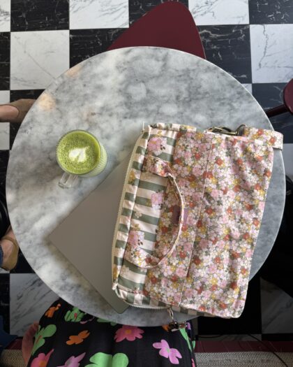 Okrągły marmurowy stolik na tle czarno-białej szachownicy, na stoliku laptop podłożony pod kolorową, kwiatową torbą i filiżanka zielonej herbaty latte z pianką w kształcie serca, widoczne fragmenty kolorowej sukienki użytkownika i obuwia.